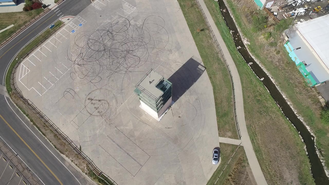 High aerial frame over Hidden Lake, Colorado showing an empty concrete lot, dark tire marks, a small building with a long shadow, nearby road edges, grass, and no visible people.