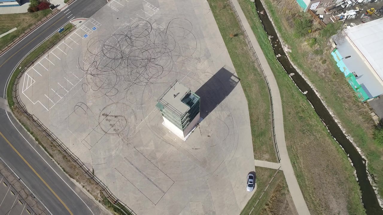 A later aerial frame in Hidden Lake, Colorado keeps the tire-marked pavement centered while the curved road, grass verge, drainage channel, small building, and no visible people remain in view.