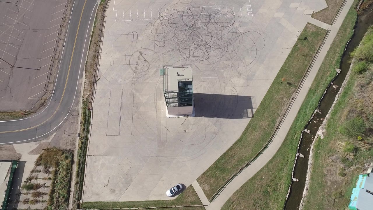 Top-down frame from Hidden Lake, Colorado showing a pale paved parking area with dark tire circles, a central tall building, a curved road at left, grass and a narrow channel at right, and no visible people.