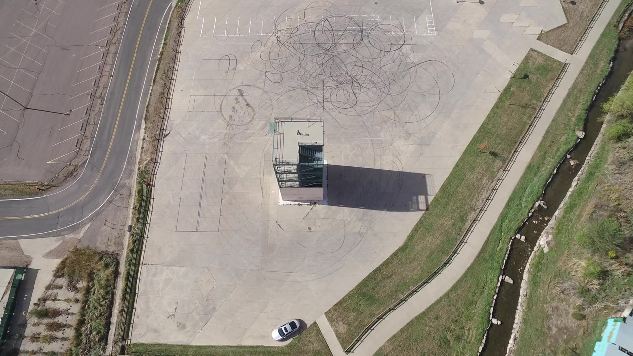 Overhead parking lot with circular tire marks, building, road, sidewalk, channel, and parked white car.