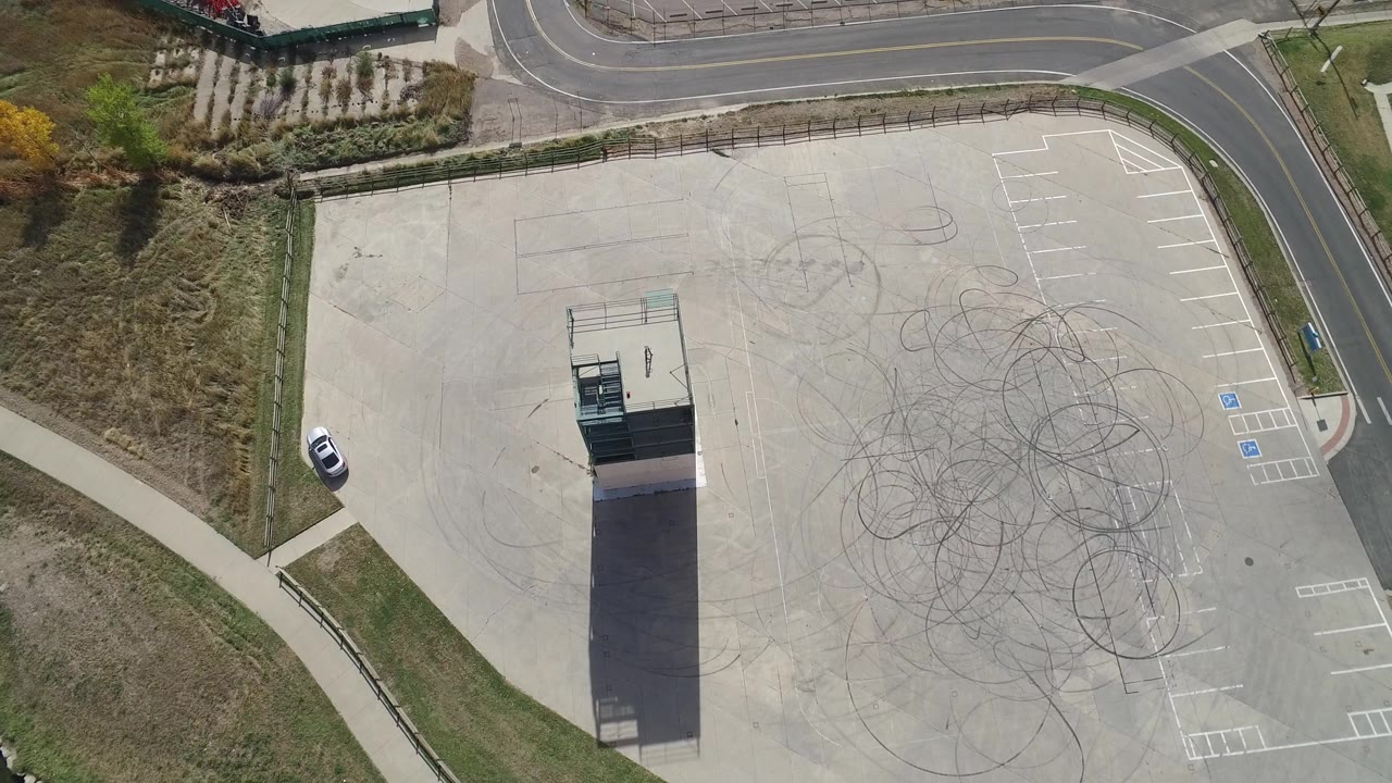 At 0:01, the Hidden Lake, Colorado aerial view keeps the tower near center, with circular tire marks spread across the concrete, empty parking spaces on the right, roadside grass, and no visible people.