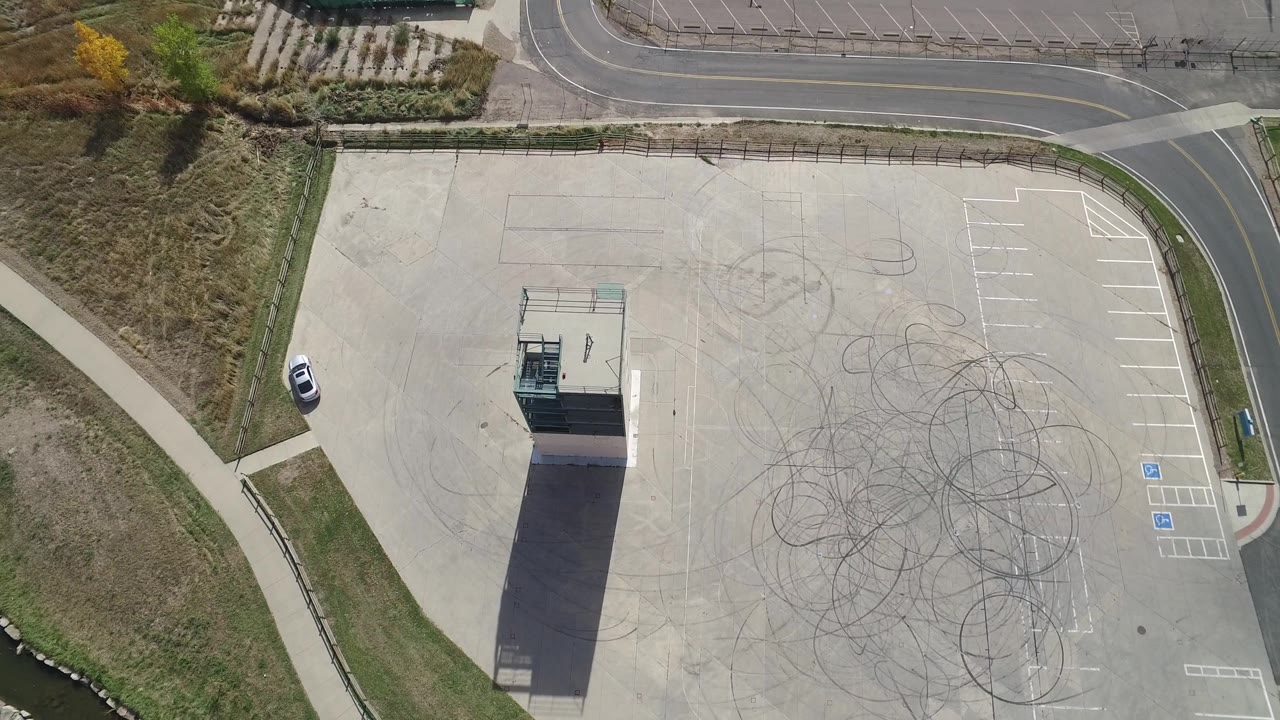 Top-down drone view of a paved lot, tower building, road, and grass drainage edge.