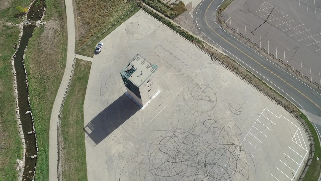 Top-down aerial frame from Hidden Lake, Colorado showing a small tower on a pale paved lot, dark tire-mark scribbles, a curved road, creek channel, grass, and no visible people.