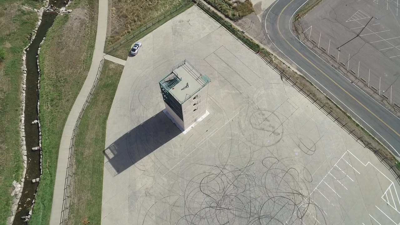Overhead frame in Hidden Lake, Colorado showing a concrete tower casting a long shadow across a pale pad with tire skid circles, grass, a road, and no visible people.