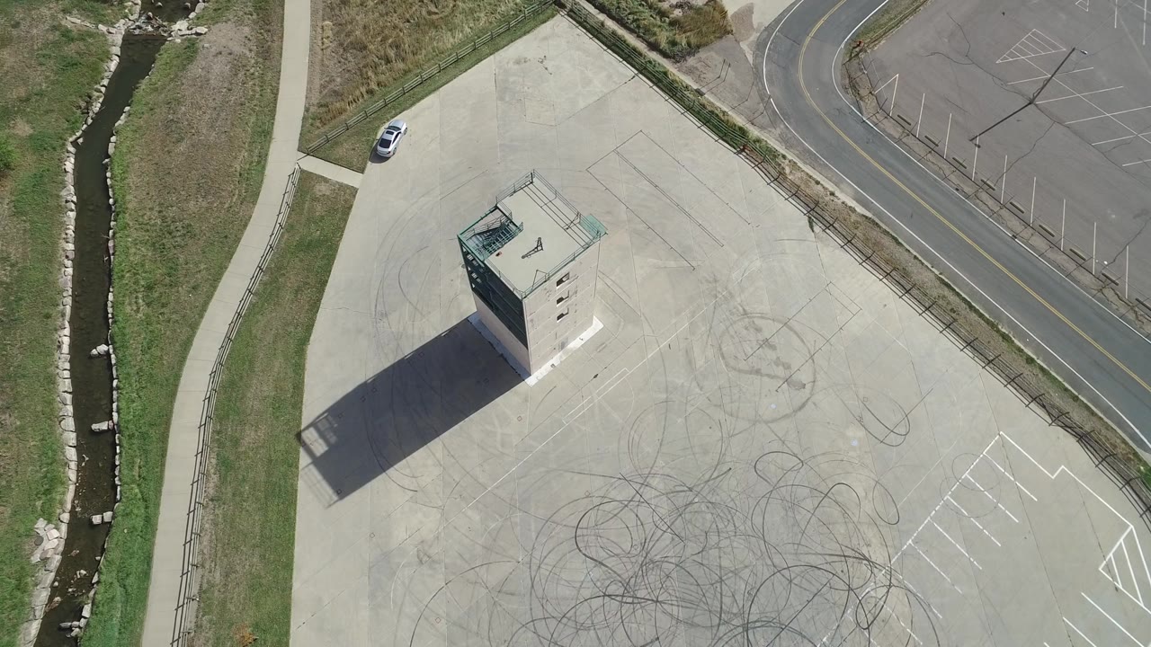 Aerial frame in Hidden Lake, Colorado with the concrete tower near the left side of a broad paved area, circular skid marks below it, a roadside edge, and no visible people.