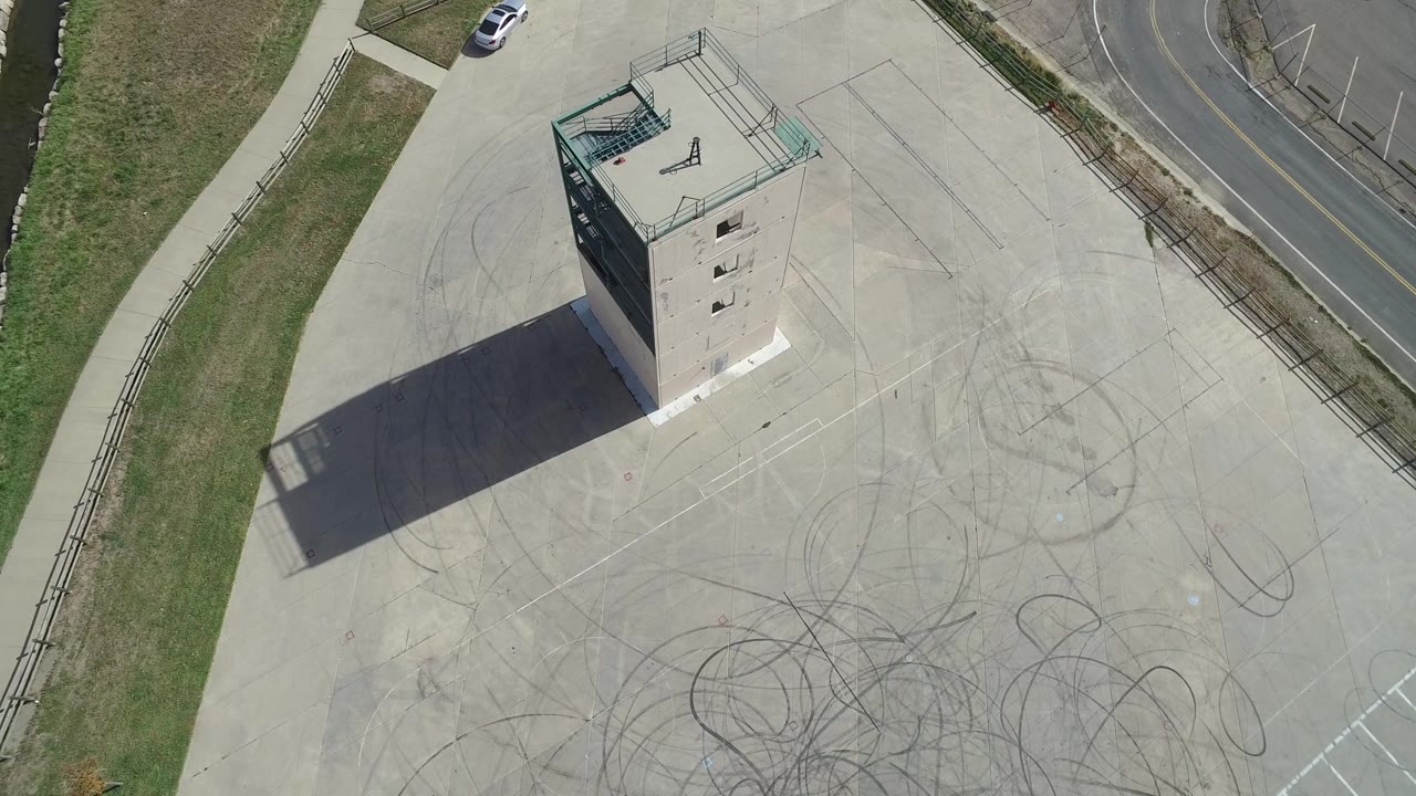 High overhead view in Hidden Lake, Colorado with the tower slightly left of center, faint circular tire marks on the concrete, roadside grass, a white car near the road, and no visible people.