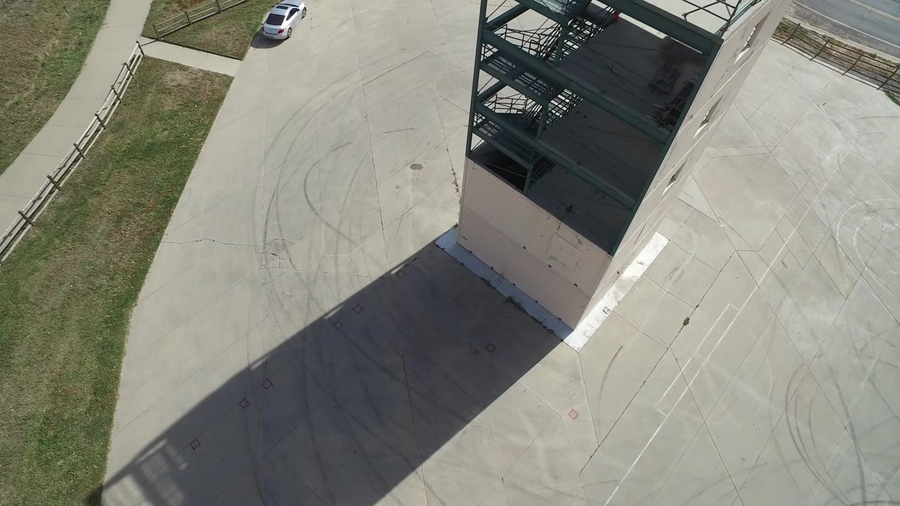 Opening overhead frame in Hidden Lake, Colorado, showing a white car near the upper edge, a fenced grass strip, a tall building, its long shadow across concrete, and no visible people.