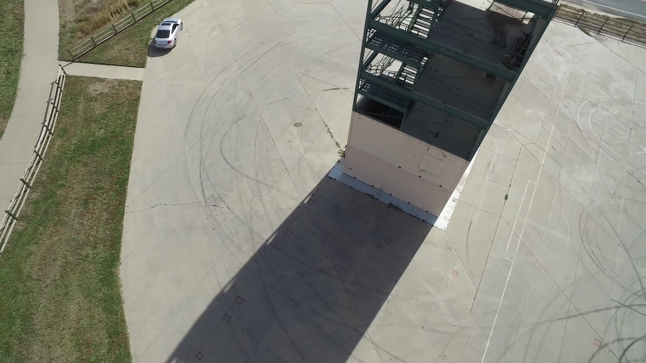 Top-down frame with white car, grass verge, building, long shadow, and marked concrete.