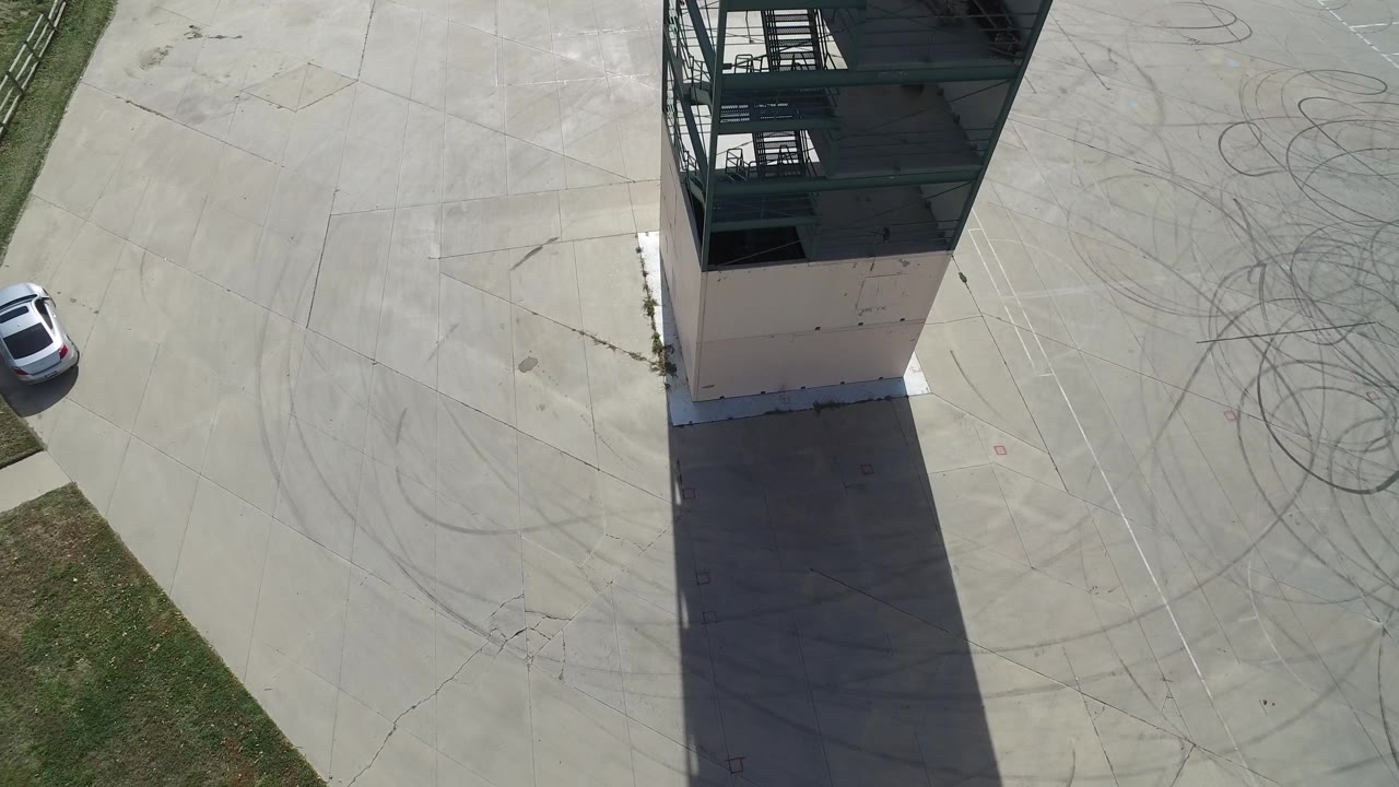 Hidden Lake, Colorado aerial frame with the glass tower near the top center, a long rectangular shadow across concrete, looping tire marks to the right, grass along the lower right, and no visible people.