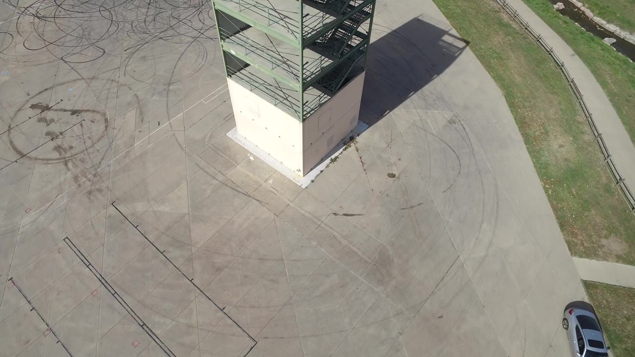 Overhead Hidden Lake, Colorado view with the tower base shifted near the upper center, looping tire marks across the concrete, grass to the right, a partial silver car, and no visible people.