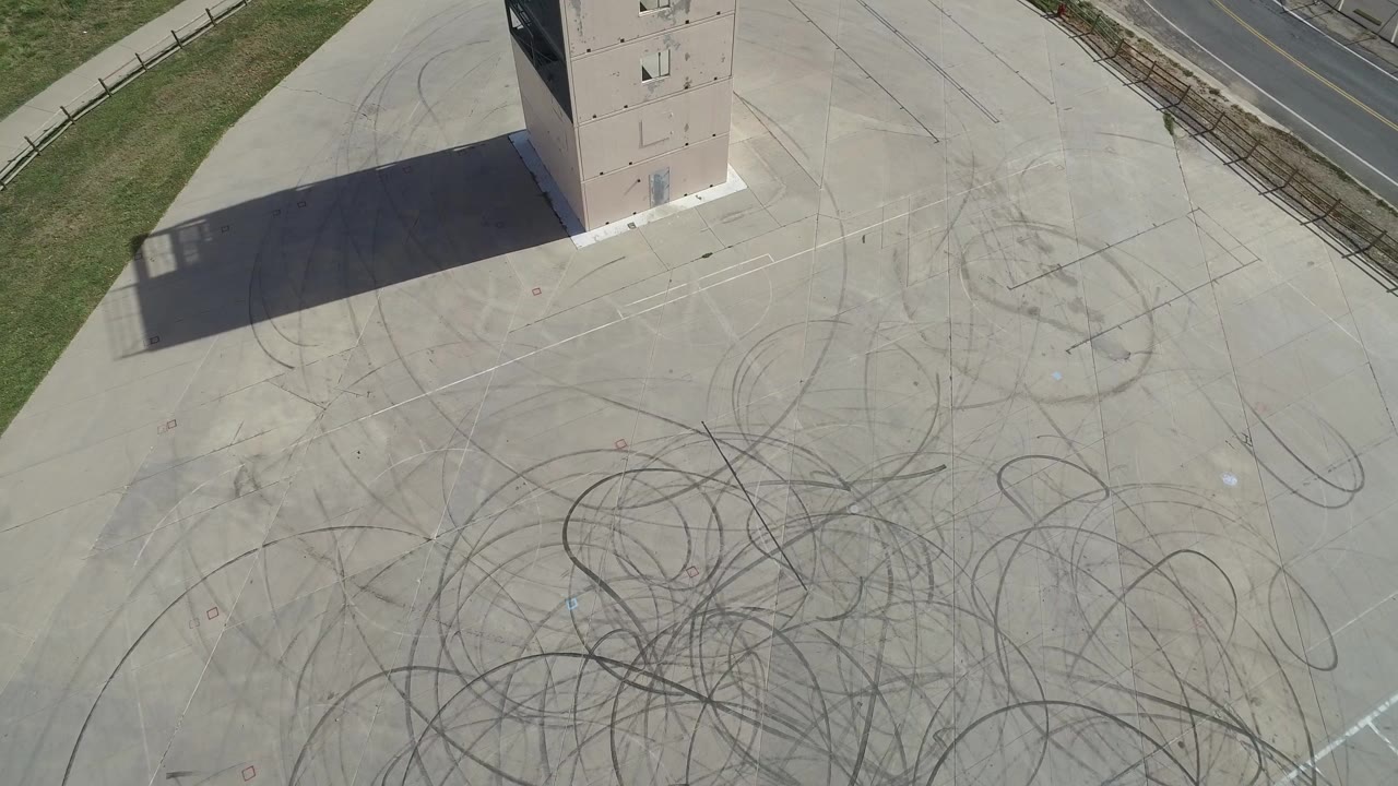 Top-down aerial frame from Hidden Lake, Colorado showing a pale concrete lot, dark looping tire marks, the base of a tower near the top edge, a long shadow, and no visible people.