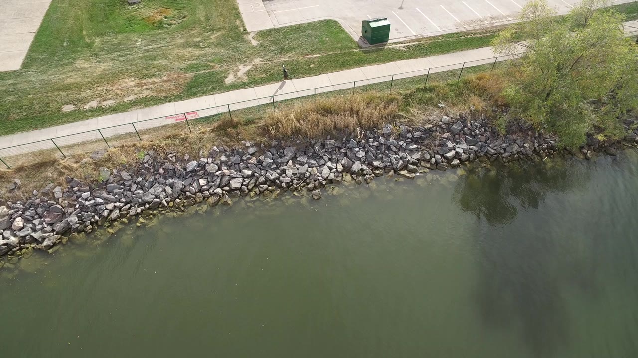 Aerial frame from Hidden Lake, Colorado showing green lake water below a rocky bank, grassy strip, paved path, fence, and no visible people.