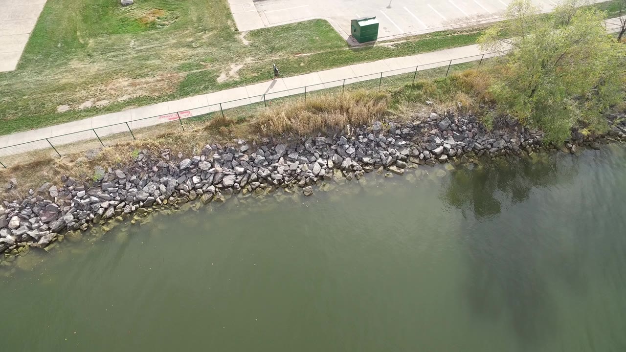 Overhead lake edge with rocks, grass, fence, and green water.