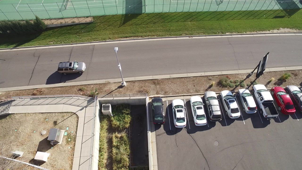 High overhead frame from Hidden Lake, Colorado showing a sunlit street, a small light SUV at left, packed parked cars below, green tennis courts above, and no clearly visible people.