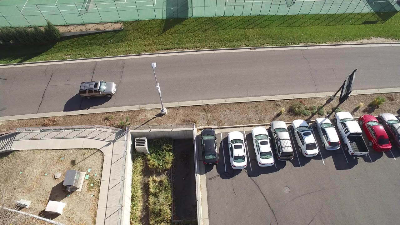 Aerial Hidden Lake, Colorado frame with the light SUV farther along the road, parked cars lined below the curb, tennis courts at the top, sidewalk paths, and no clearly visible people.