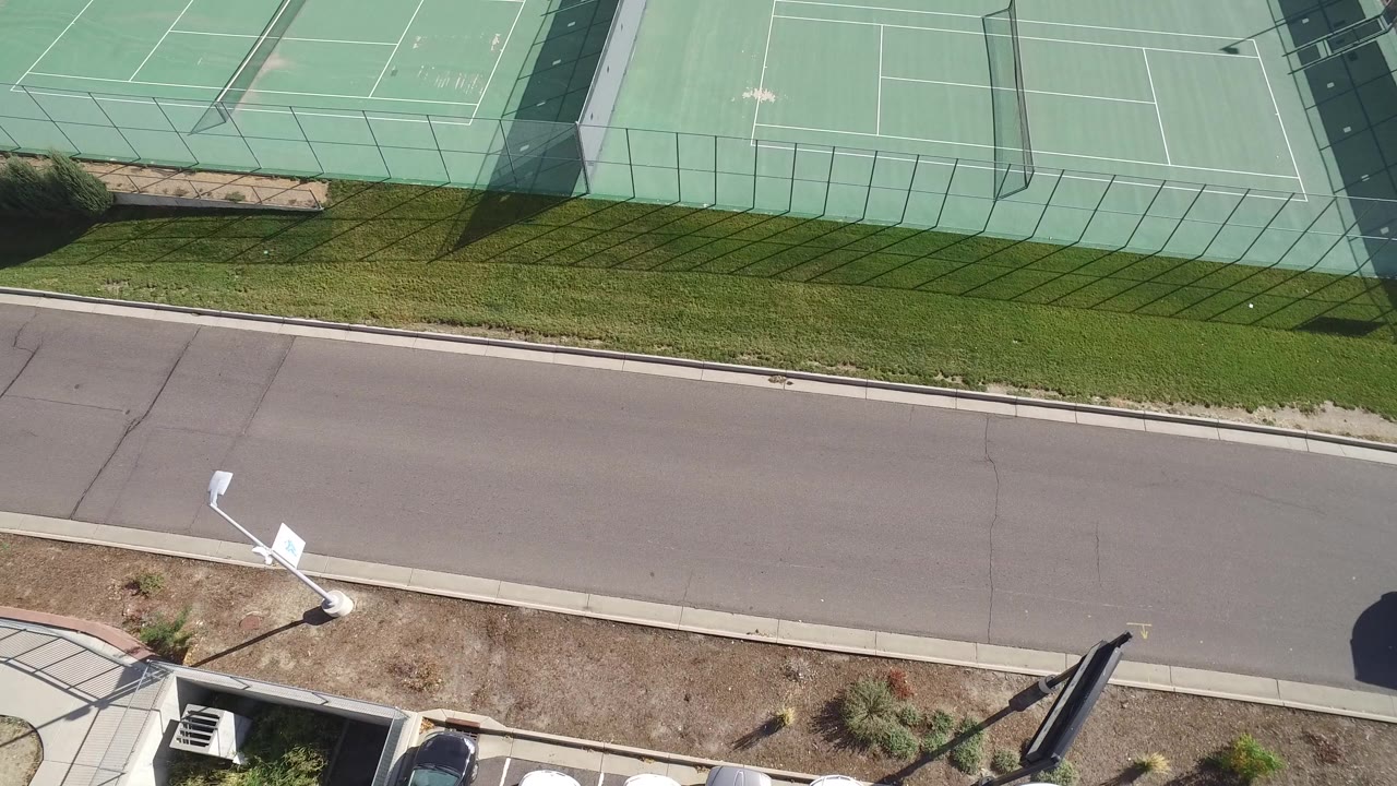 Overhead frame in Hidden Lake, Colorado showing green tennis courts, fencing, a road, grass, light poles, and no visible people at the start of the clip.