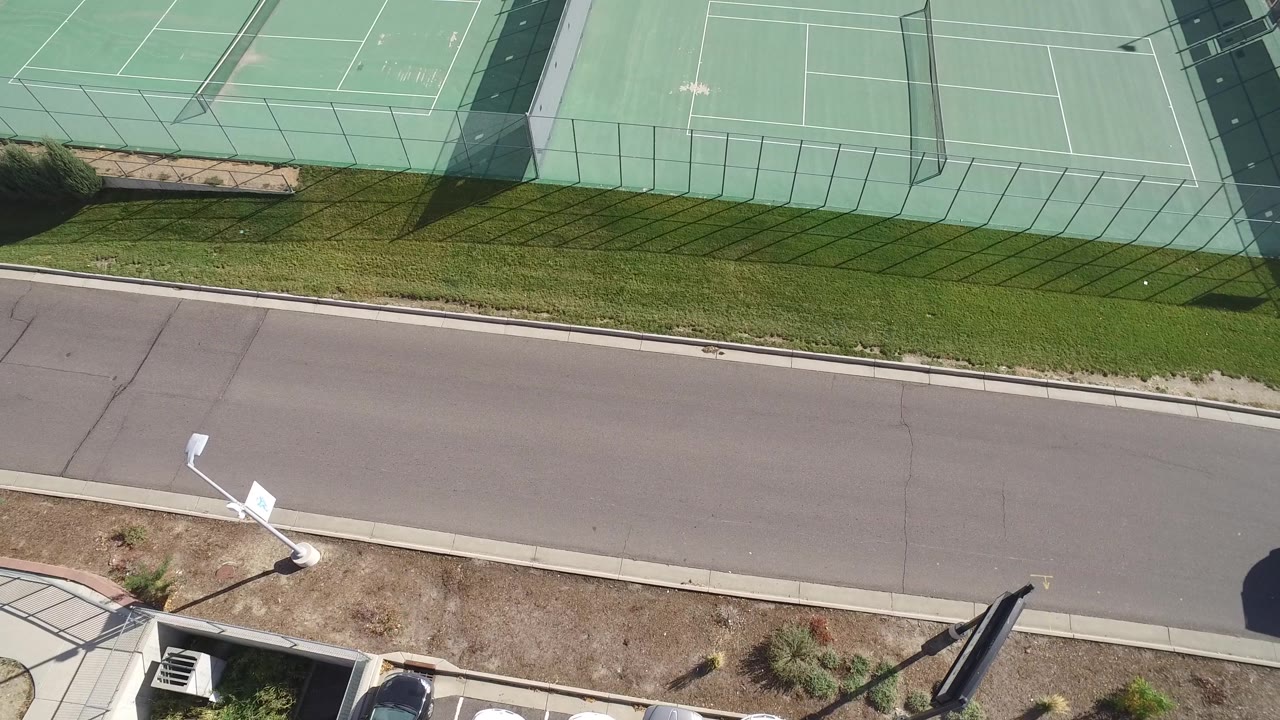 Aerial frame in Hidden Lake, Colorado with fenced green tennis courts at the top, an empty paved road below, light poles near the curb, and no visible people.
