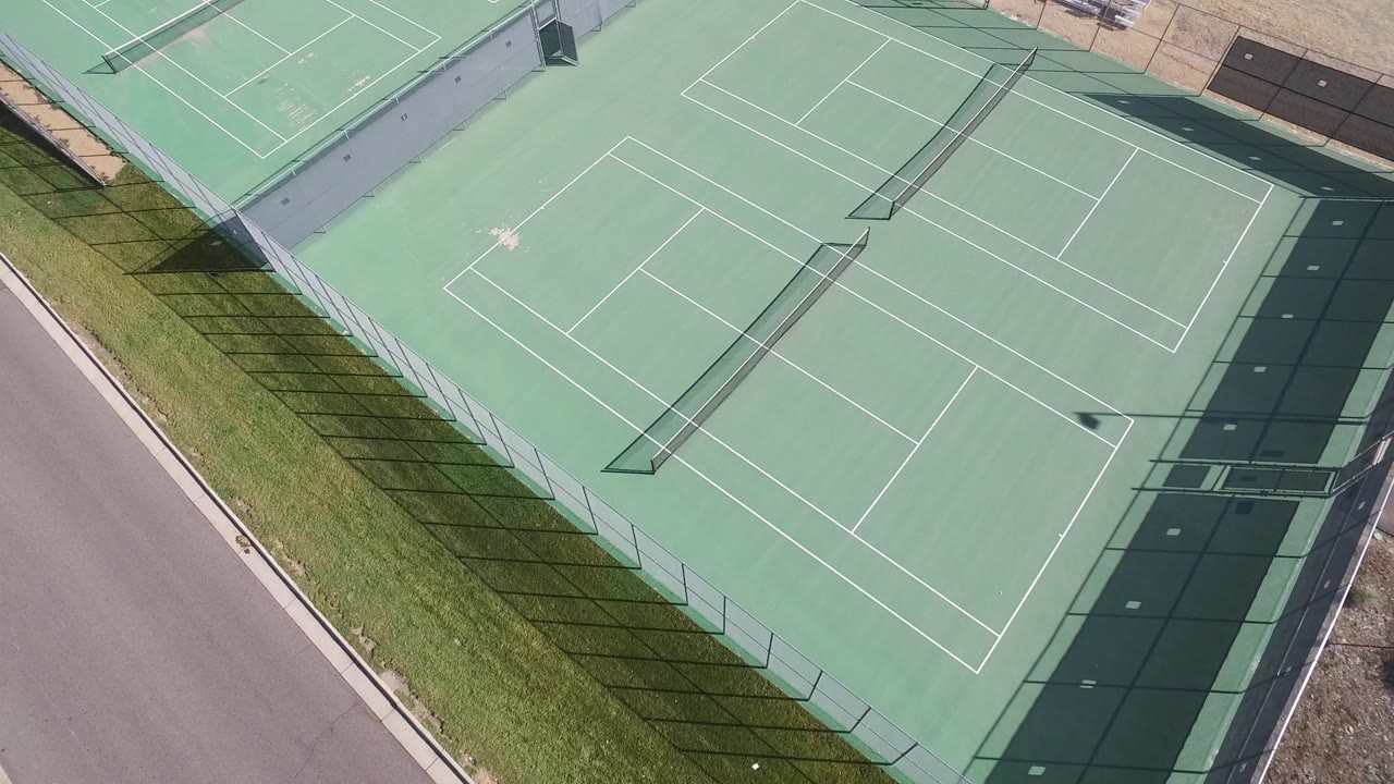 Overhead view of the Hidden Lake tennis court with the court surface centered, long fence shadows crossing the left grass strip, the road along the lower edge, and no visible people.