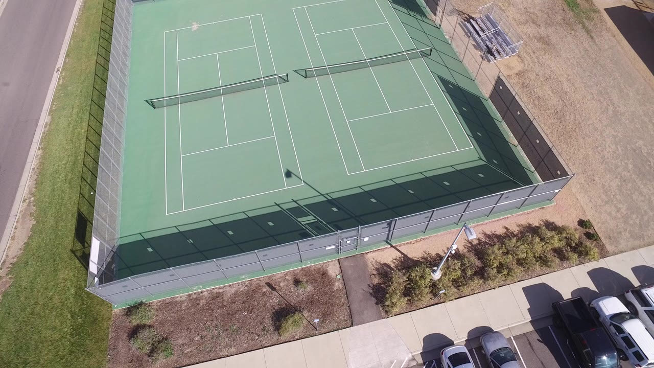 Overhead view of fenced green tennis courts beside a sidewalk and parked cars.