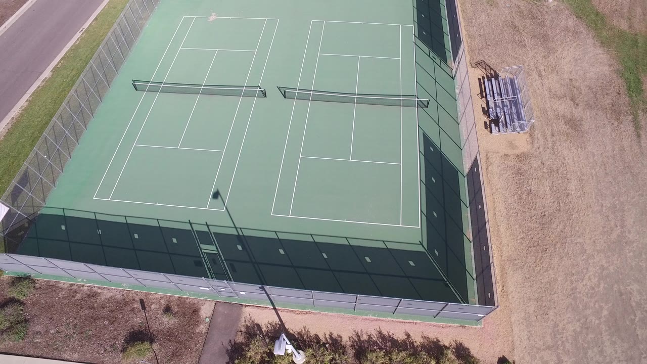 Top-down frame over two green fenced tennis courts in Hidden Lake, Colorado, with dry tan ground, a road edge, and no visible people.