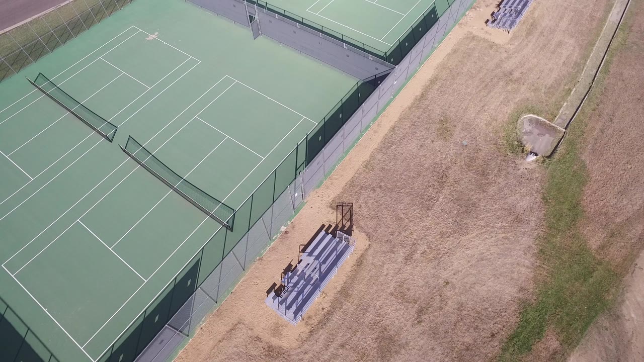 Aerial view of green tennis courts beside dry grass and bleachers.