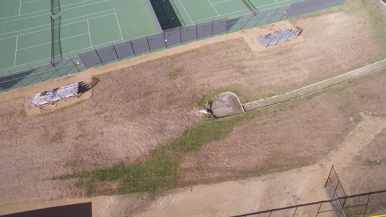 A second overhead view in Hidden Lake, Colorado keeps the fenced tennis courts along the top edge while dry grass, dirt, stacked materials, and a concrete drainage feature fill the open ground, with no visible people.