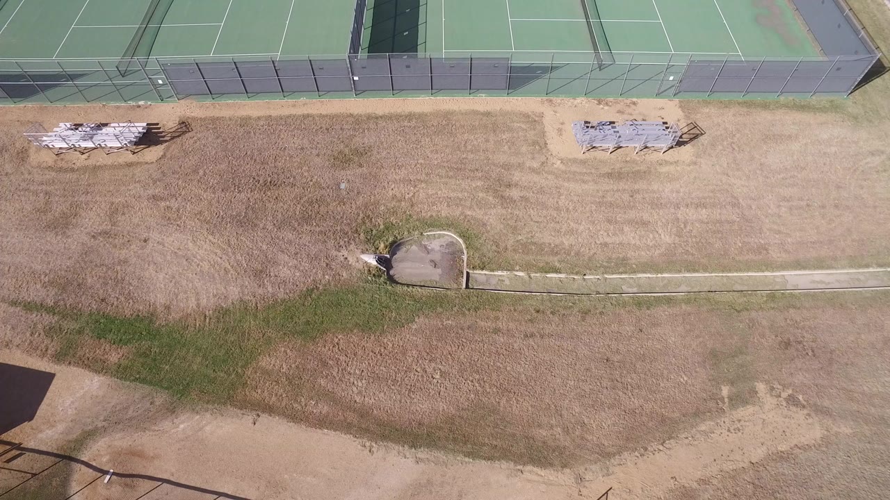 Overhead view of green tennis courts, dry grass, fencing, bleachers, and a paved path.