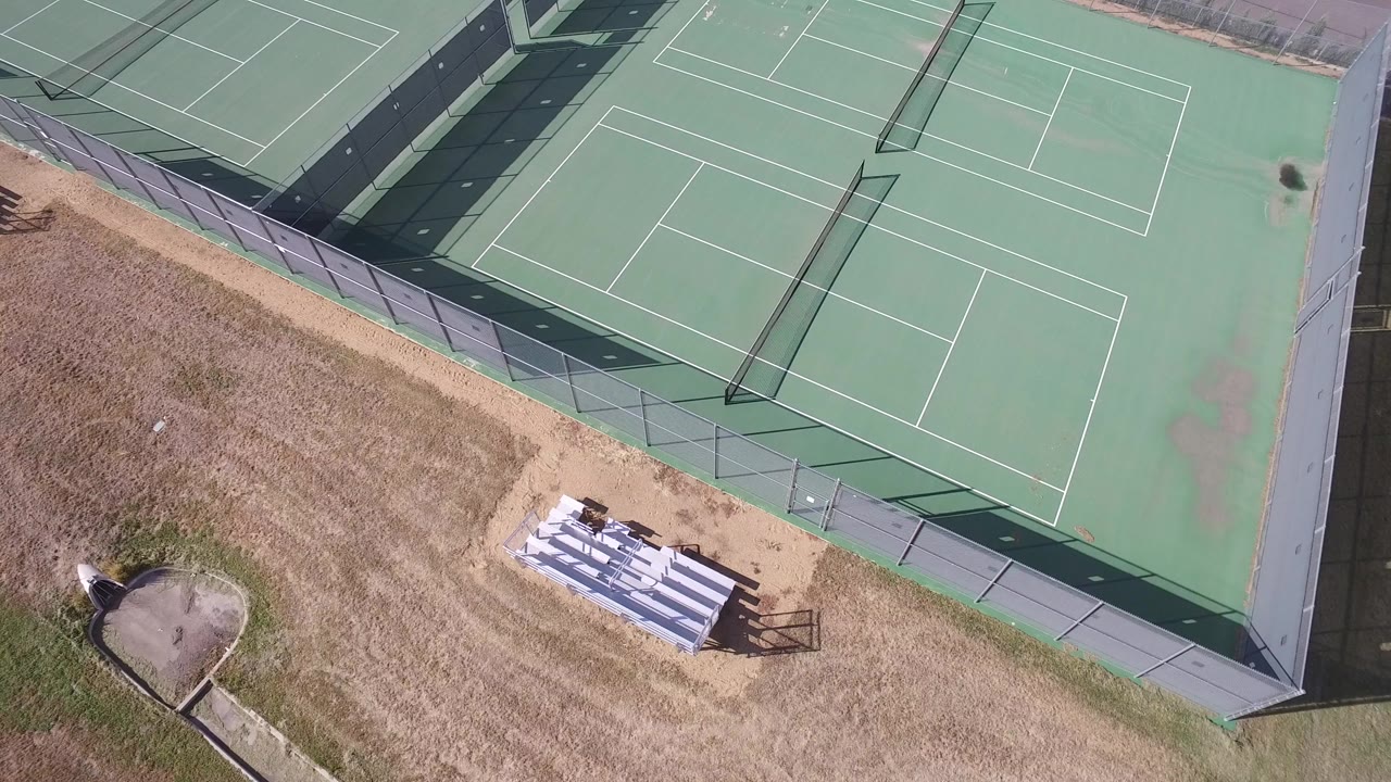 Overhead view of green tennis courts beside dry grass and white bleachers.