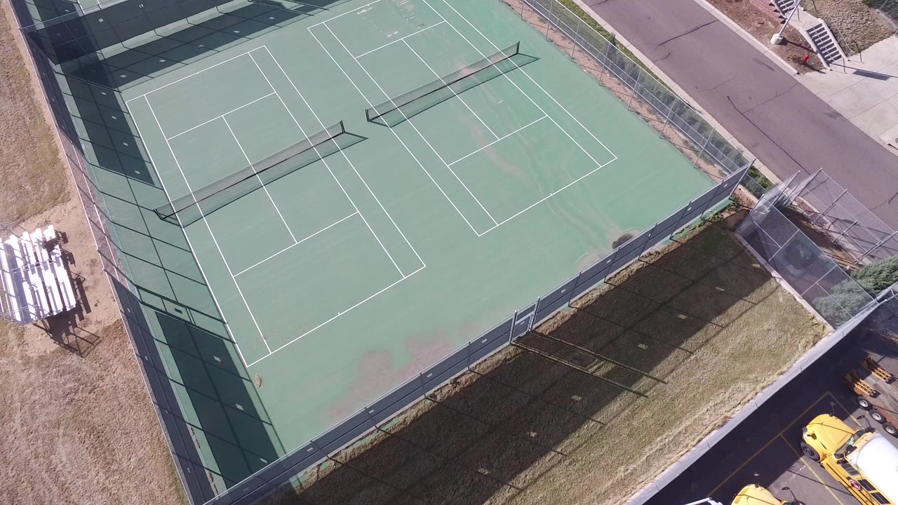 Opening overhead frame in Hidden Lake, Colorado showing two green fenced sports courts, white court lines, long net shadows, nearby pavement, and no visible people.