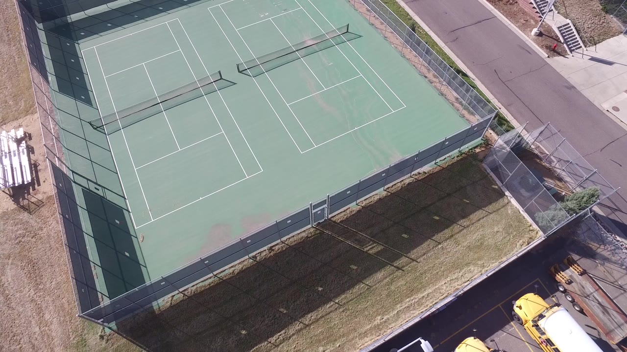 High-angle view of the fenced court corner, road, grass, and yellow vehicles below.