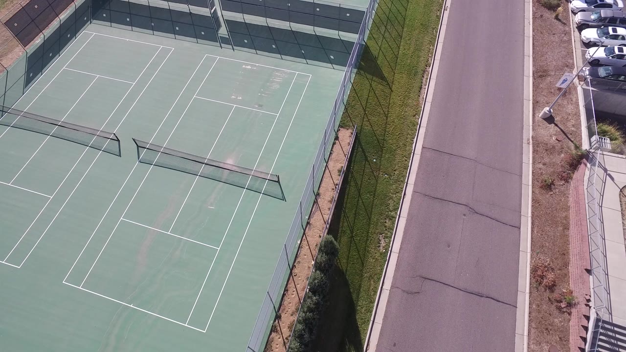 Opening overhead frame in Hidden Lake, Colorado showing green tennis courts, court lines, a tall fence, a paved road, and no visible people.