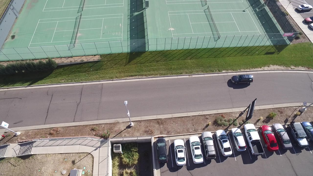 Overhead view of fenced green tennis courts beside a road and parking lot.