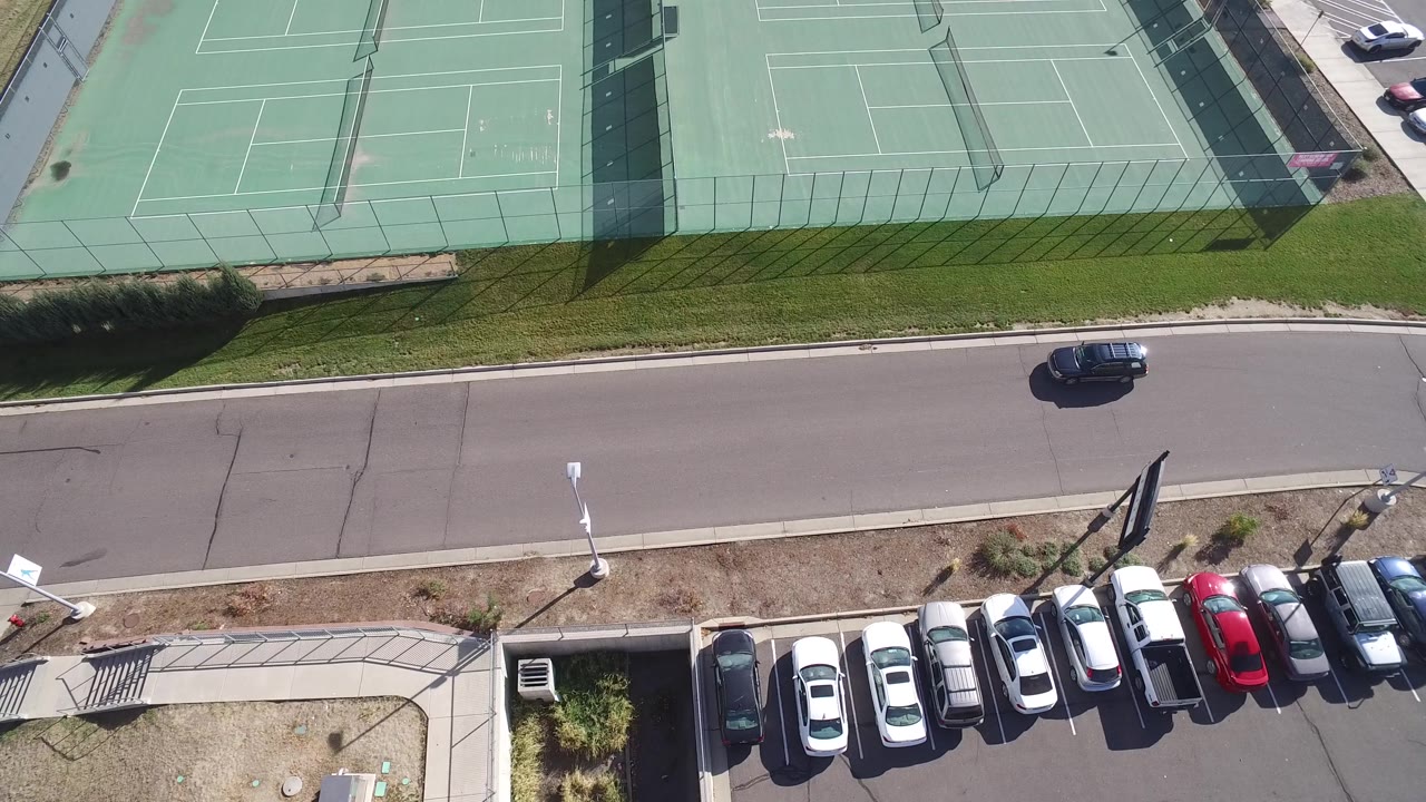 Aerial frame of the Hidden Lake tennis courts with a dark car on the road, rows of parked vehicles, sidewalks, fencing, and small people visible on the courts.