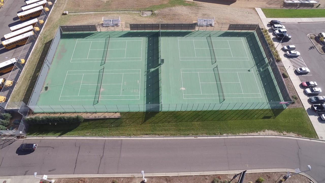 Overhead view of green tennis courts beside a road and parked cars.