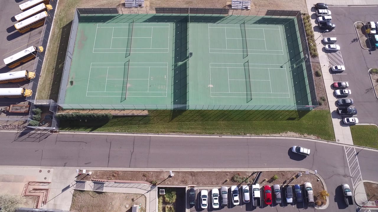 Overhead view of fenced tennis courts beside a road and parking areas.