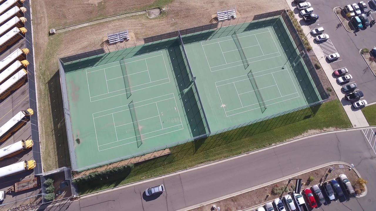 Overhead frame near Hidden Lake, Colorado showing two fenced green tennis courts, a curved road, parked cars, dry grass, and no visible people.