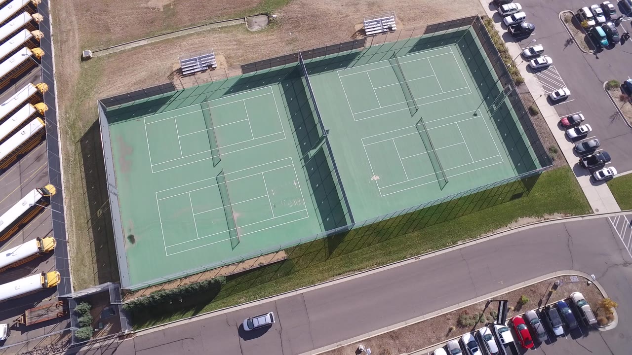 Second overhead frame near Hidden Lake, Colorado holding on the fenced tennis courts, surrounding dry grass, parked cars along the lot, and no visible people.