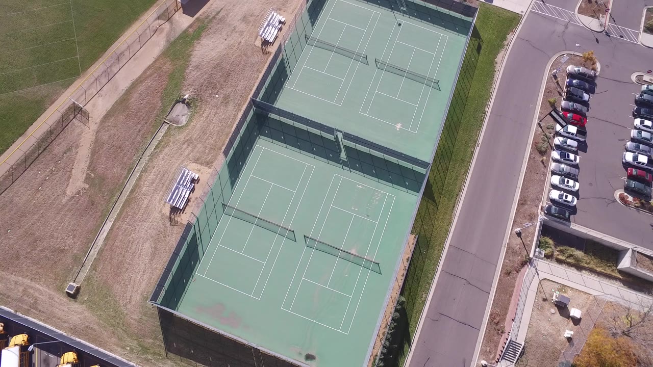 Aerial view of two empty fenced tennis courts beside a road and parking lot.