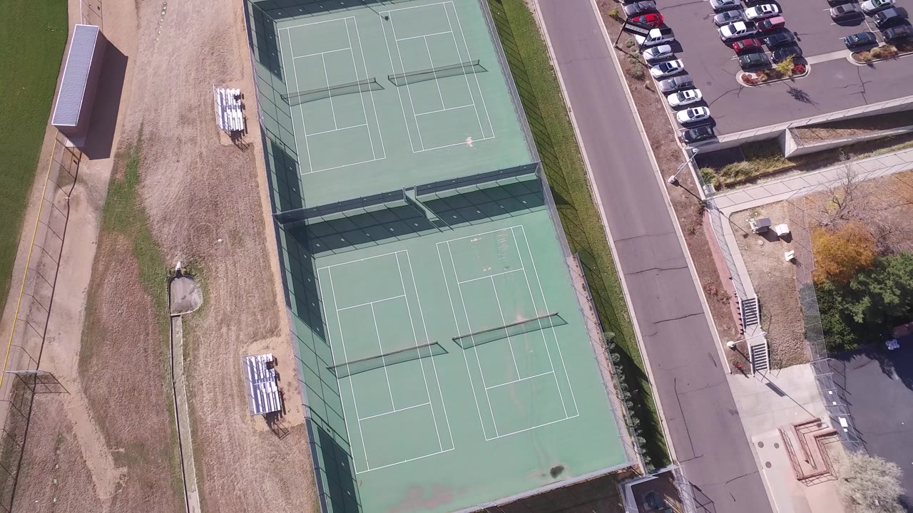 Overhead frame from Hidden Lake, Colorado showing two fenced green tennis courts beside dry grass, a road, parked cars, bleachers, and no visible people.