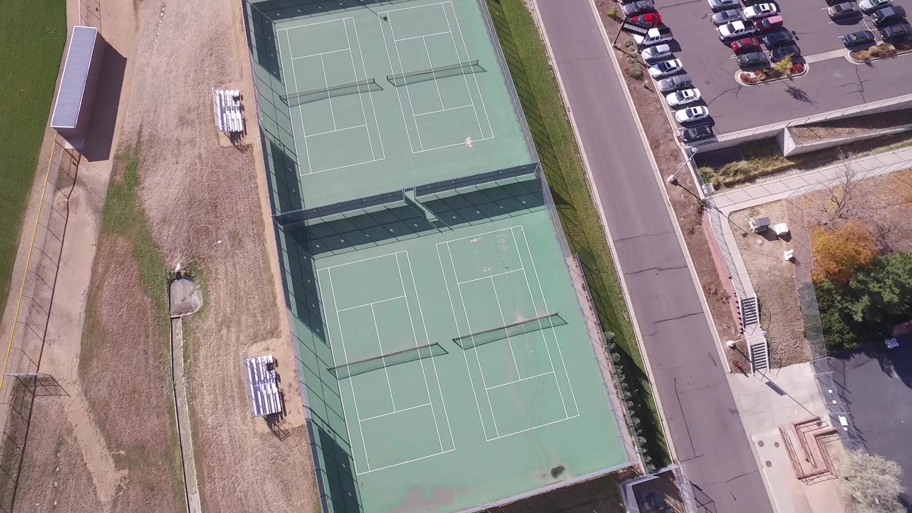 Top-down Hidden Lake, Colorado view with the paired tennis courts centered between a dry grass strip, adjacent street, parked vehicles, and no visible people.