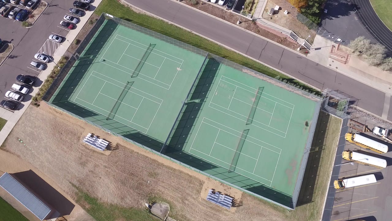 Overhead frame in Hidden Lake, Colorado showing two green fenced tennis court areas, dry grass, parked cars, road edges, and no visible people.