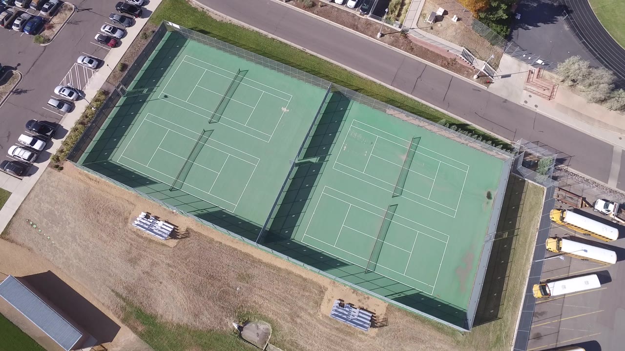 Aerial frame in Hidden Lake, Colorado with the tennis courts centered, a row of parked cars at the upper road, dry grass below, and no visible people.