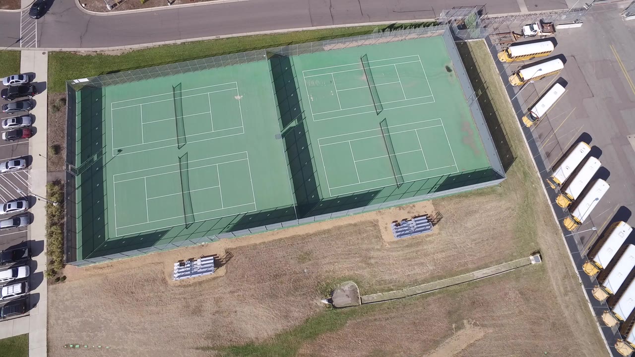 Overhead view of two fenced tennis courts beside parking and dry grass.