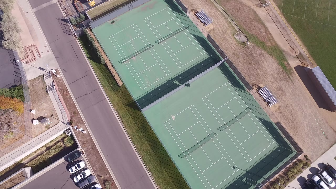 High overhead frame of two green fenced tennis courts in Hidden Lake, Colorado, bordered by a road, sidewalks, parked cars, dry grass, and no visible people.