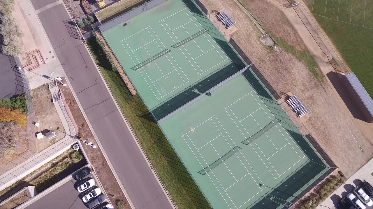 Top-down aerial view in Hidden Lake, Colorado, showing the paired tennis courts shifted slightly in frame with a road on the left, parked cars near the lower edge, and no visible people.