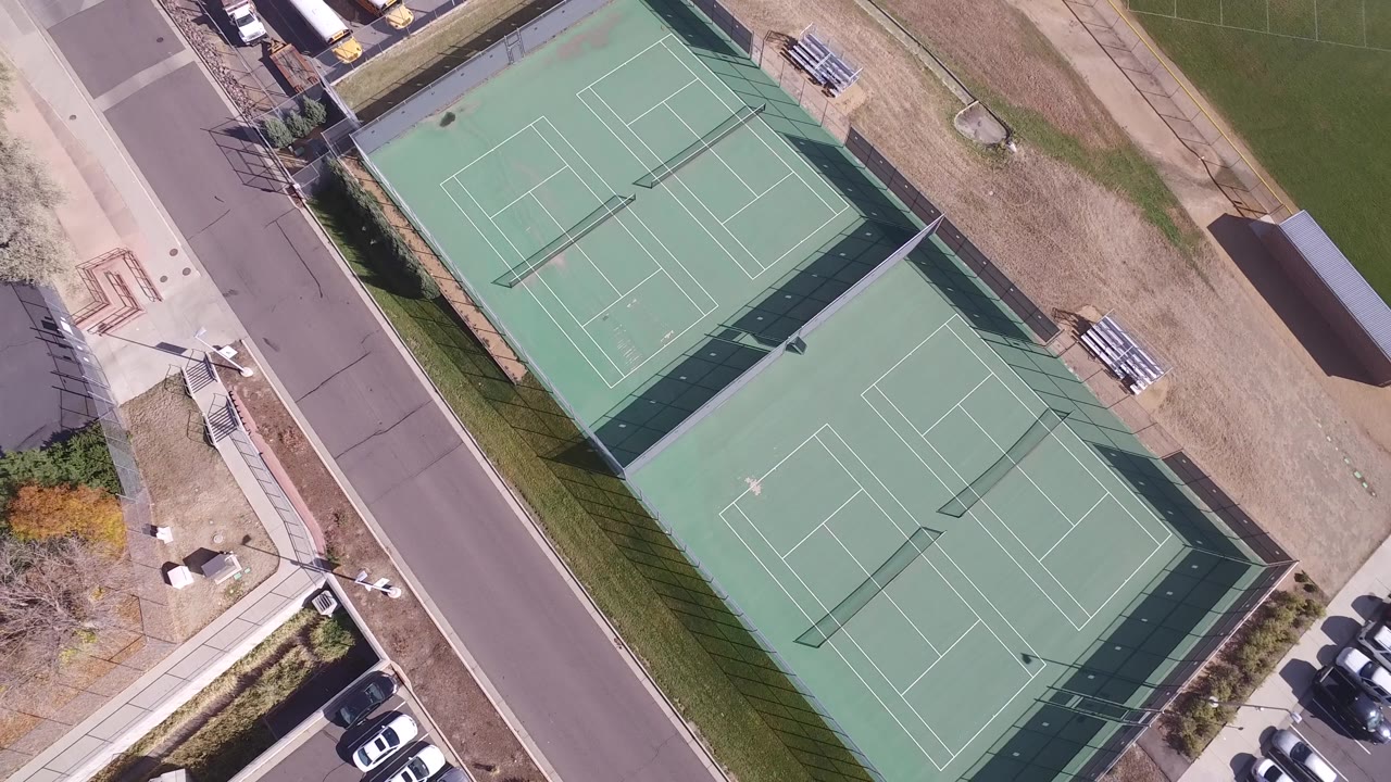 Drone view of fenced tennis courts with buses and parked cars nearby.