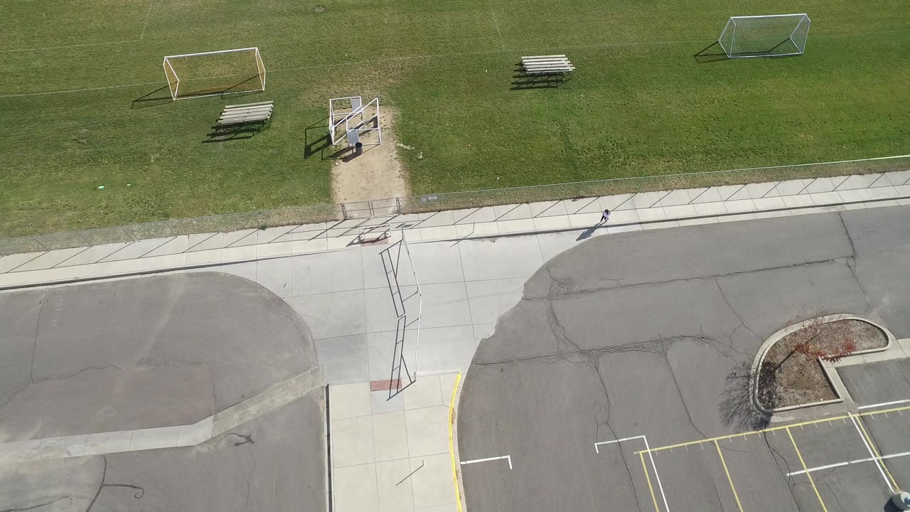 Overhead frame from Hidden Lake, Colorado showing a green sports field, two soccer goals, bleachers, sidewalk, asphalt road, and a tiny person near the right sidewalk.