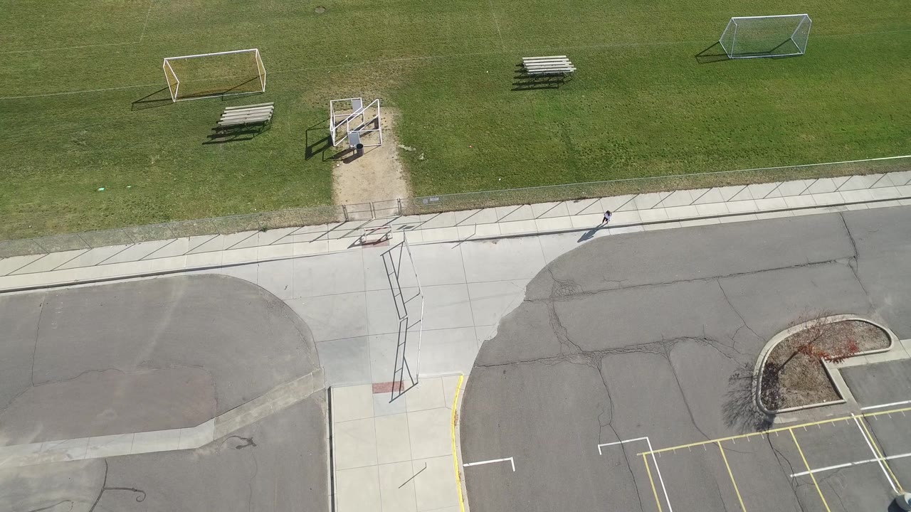 Hidden Lake, Colorado aerial still with the road curving along the bottom, a sidewalk crossing the frame, soccer goals on the grass, bleachers, and a distant pedestrian near the right edge.