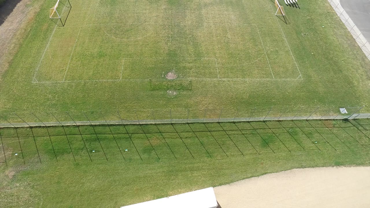 Aerial frame in Hidden Lake, Colorado with soccer field rectangles across green grass, long fence shadows through the middle, and a pale paved edge below with no visible people.