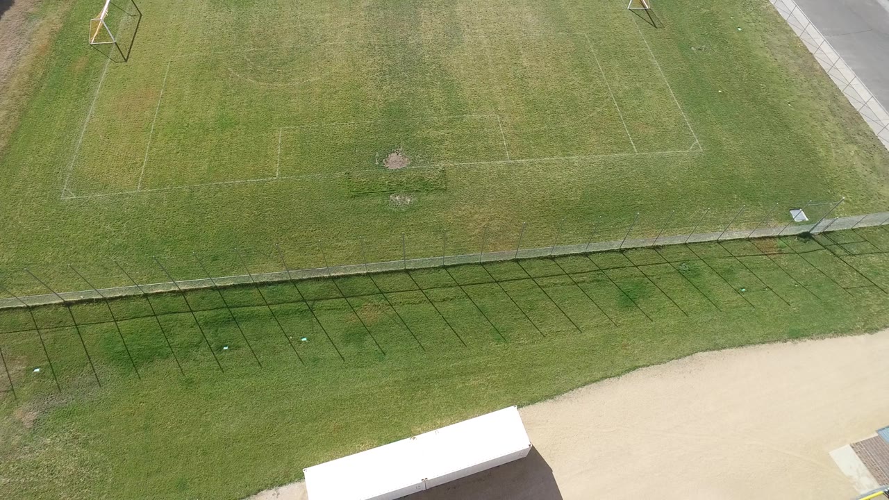 Aerial view of a soccer field, fence, pale road, and white-roofed structure edge.